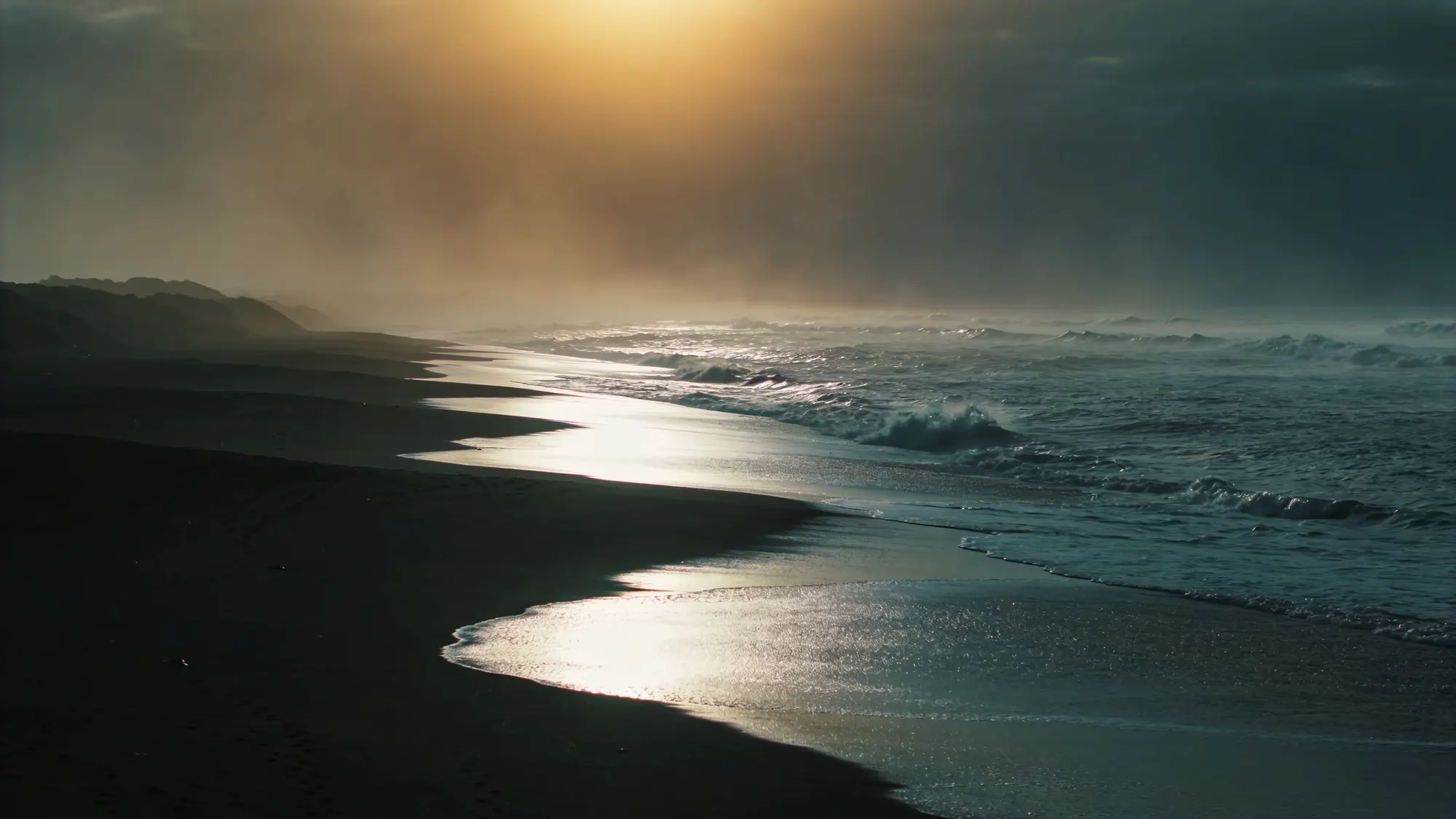 A moody New Zealand black-sand beach at first light, the tide swirling over dark sand as waves roll toward a misty headland